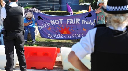 Two Quakers hold a handmade banner at the side of a road while police look on. The banner depicts fighter planes dropping bombs with a fiery destruction in the middle. It reads "This is NOT FAIR Trade! Quakers for Peace"