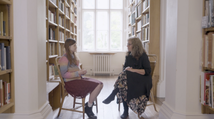 Still from the film, showing an older person and a younger person in deep conversation. They are seated in a library and look at each other as they speak.