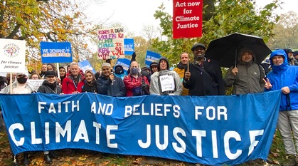 Protesters stand in the rain among trees, behind a banner reading: faith and beliefs for climate justice