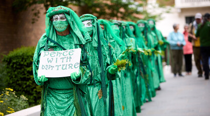 Activists dressed all in green, the front one holding a sign saying 'peace with nature' and the others holding leaves, walk in a line towards the camera