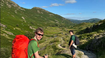 Two hikers, both with rucksacks, on a stony path high in the hills, with bright sun shining and a dramatic view of the valley